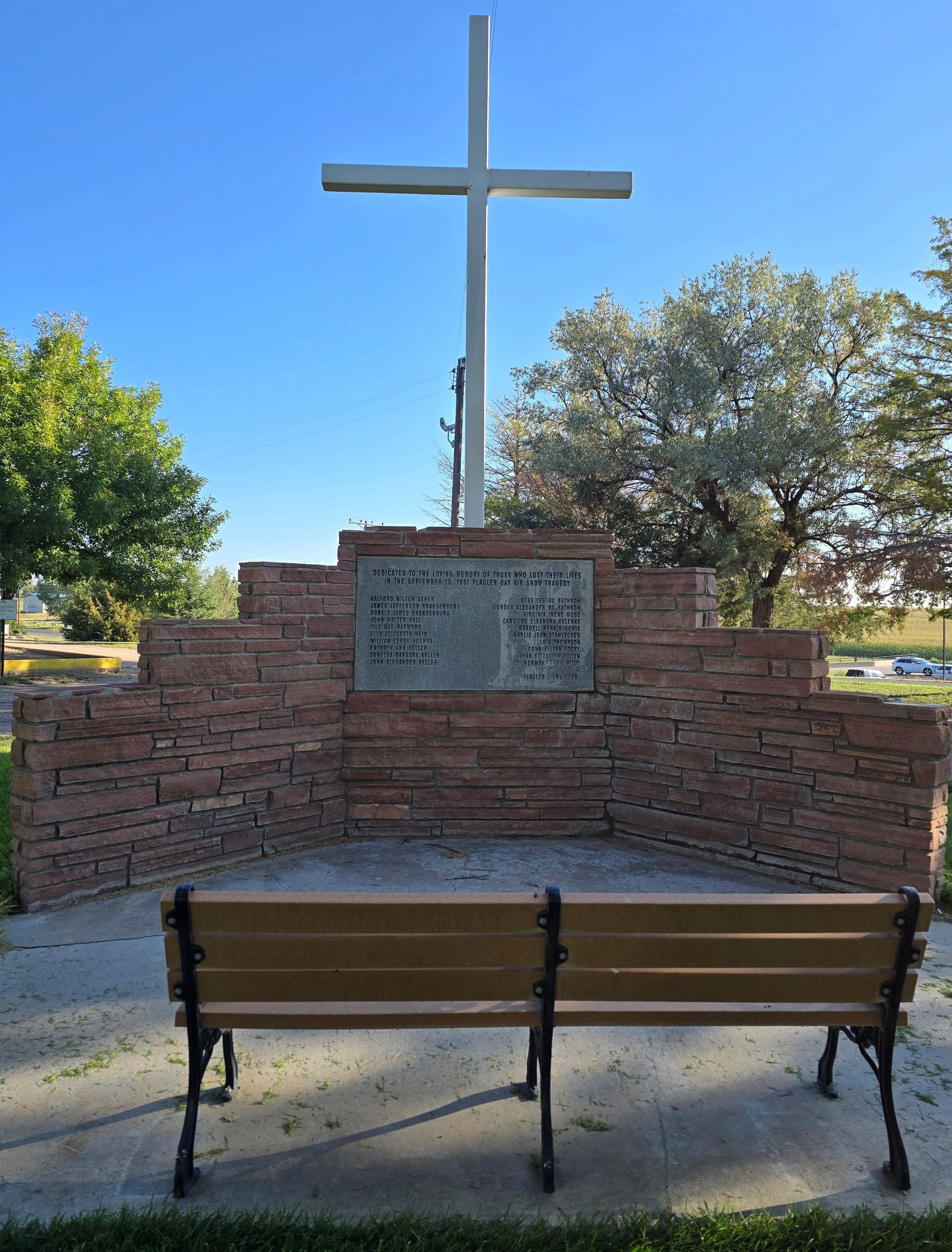 Wood bench, red brick with grey stone plaque listing names in black print, white cross