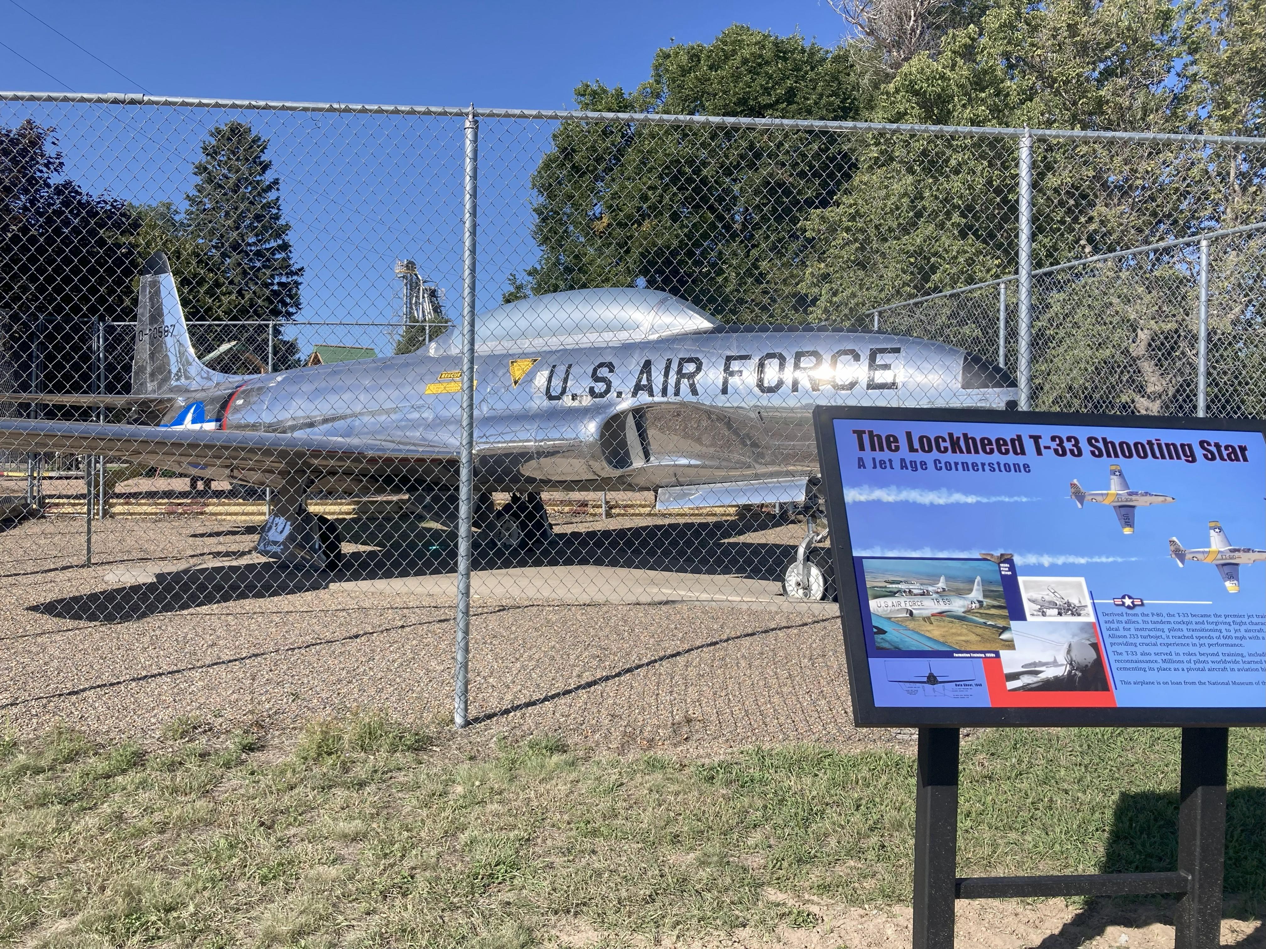 Silver jet airplane set behind chain link fence, black letters U.S. Air Force, blue sign