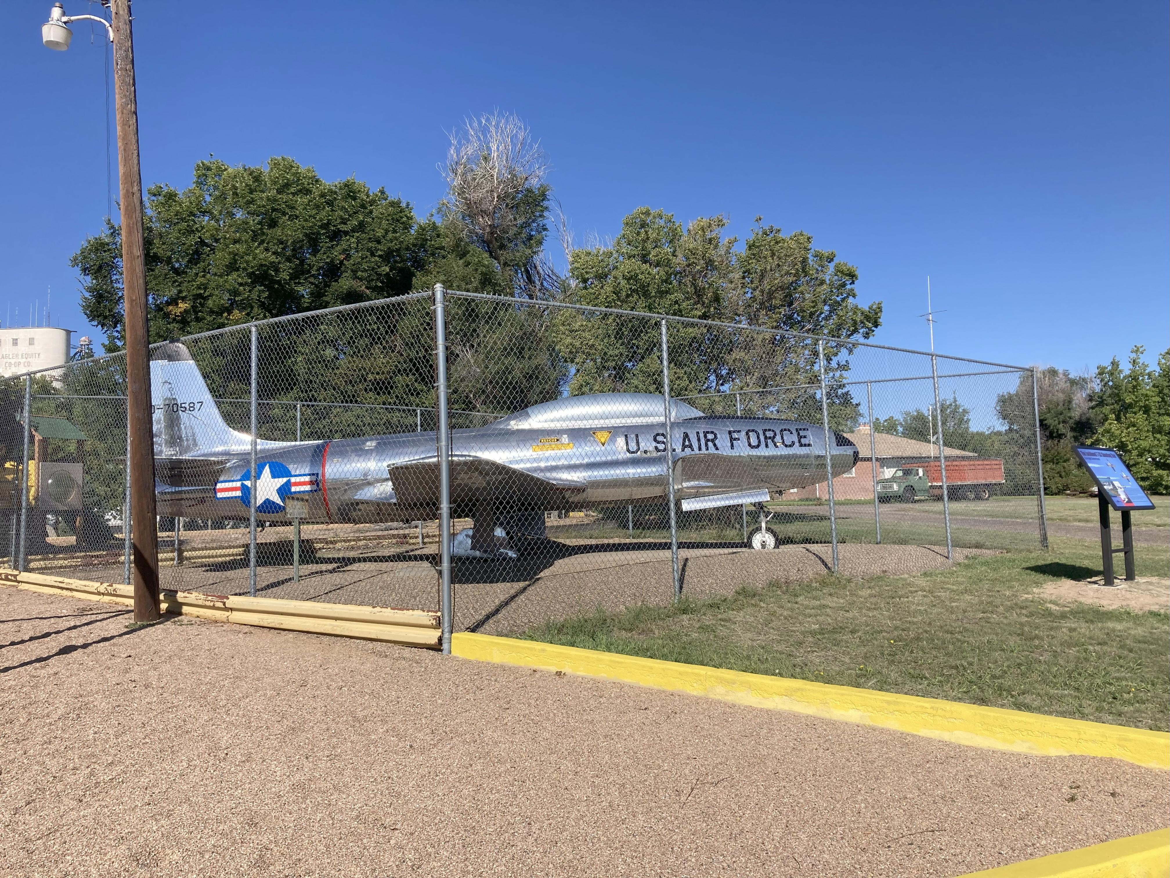 Silver jet airplane set behind chain link fence, black letters U.S. Air Force, white star emblem in circled in blue with red stripes
