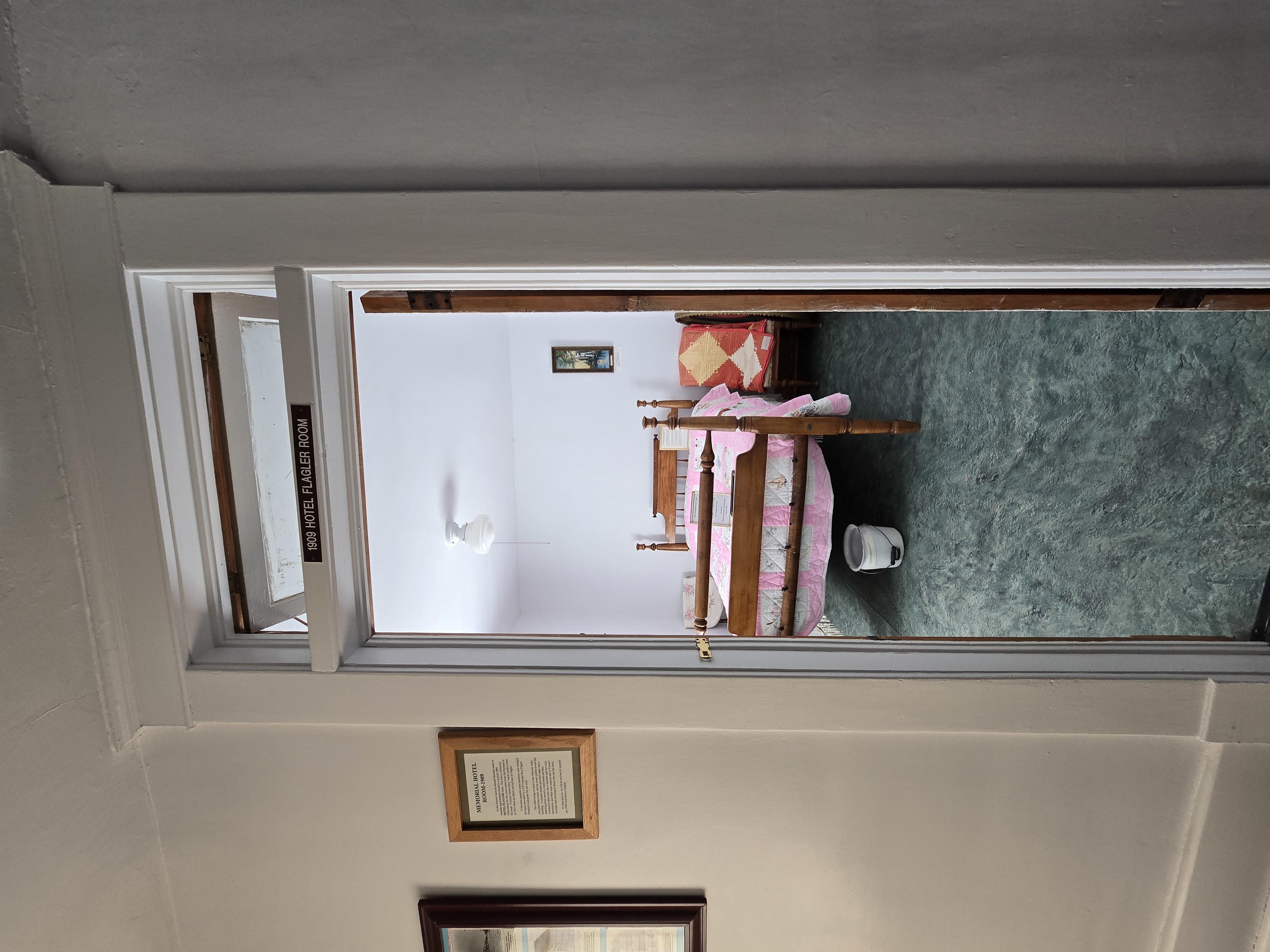 1909 Hotel Room, Wooden bed with Pink and white quilt, with white metal chamber pot with handle under bed. 