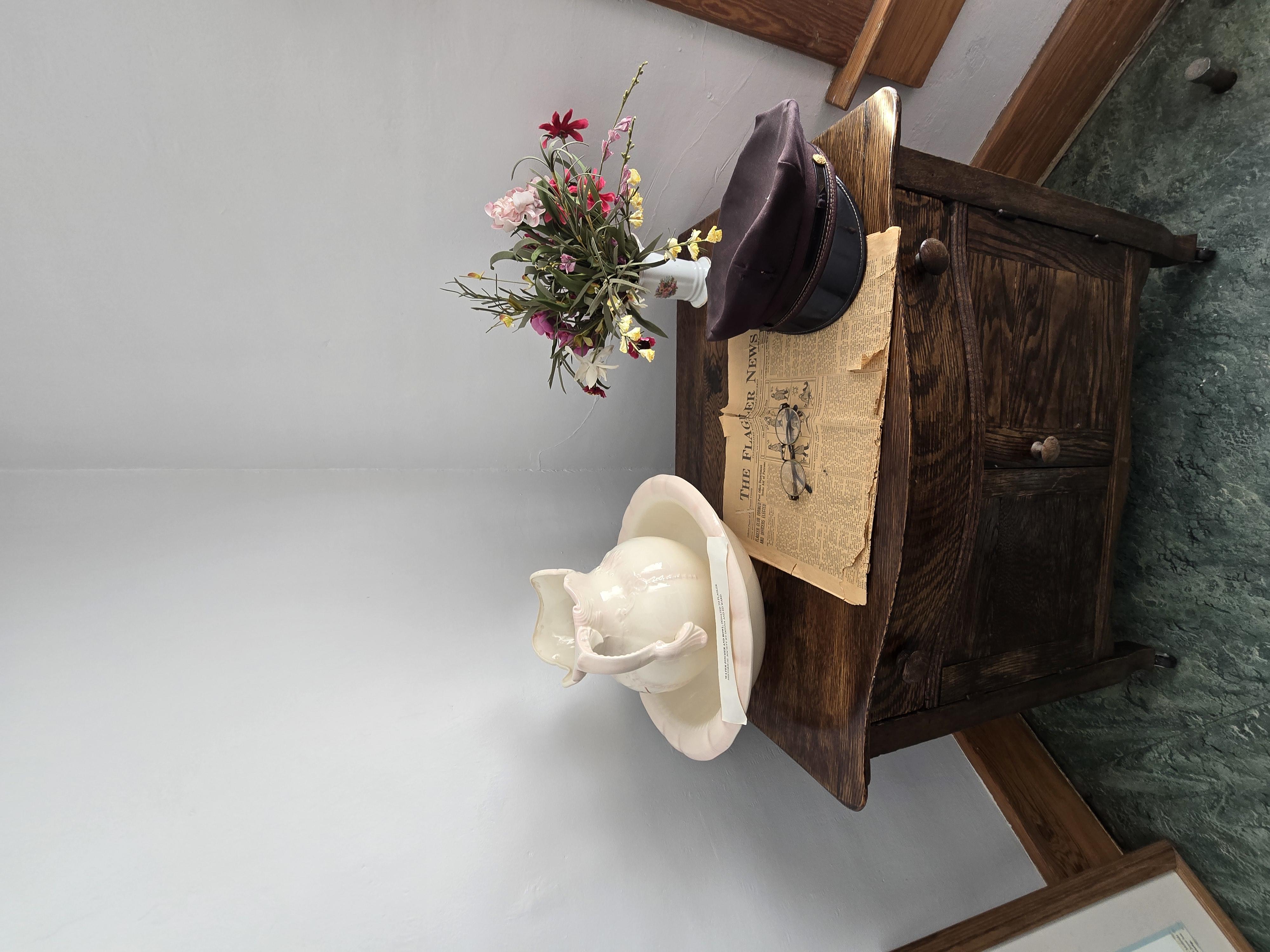White wash basin, black soldier cap, white vase of multiple color flowers, old Flagler newspaper and reading glasses on wooden cabinet