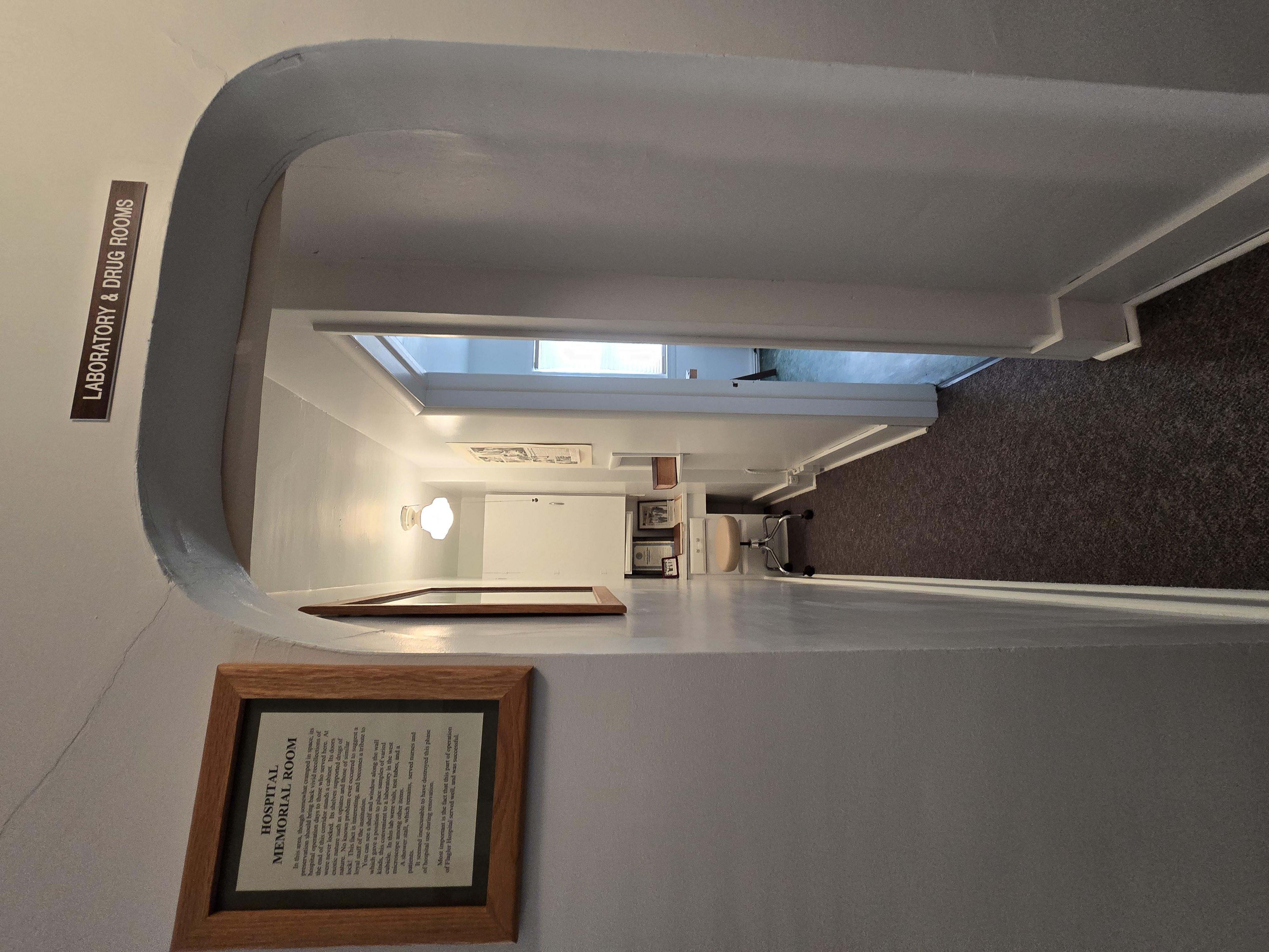 Photo of brown sign above door with white littering saying Laboratory & Drug Rooms, doctor stool, brown carpet in narrow hallway