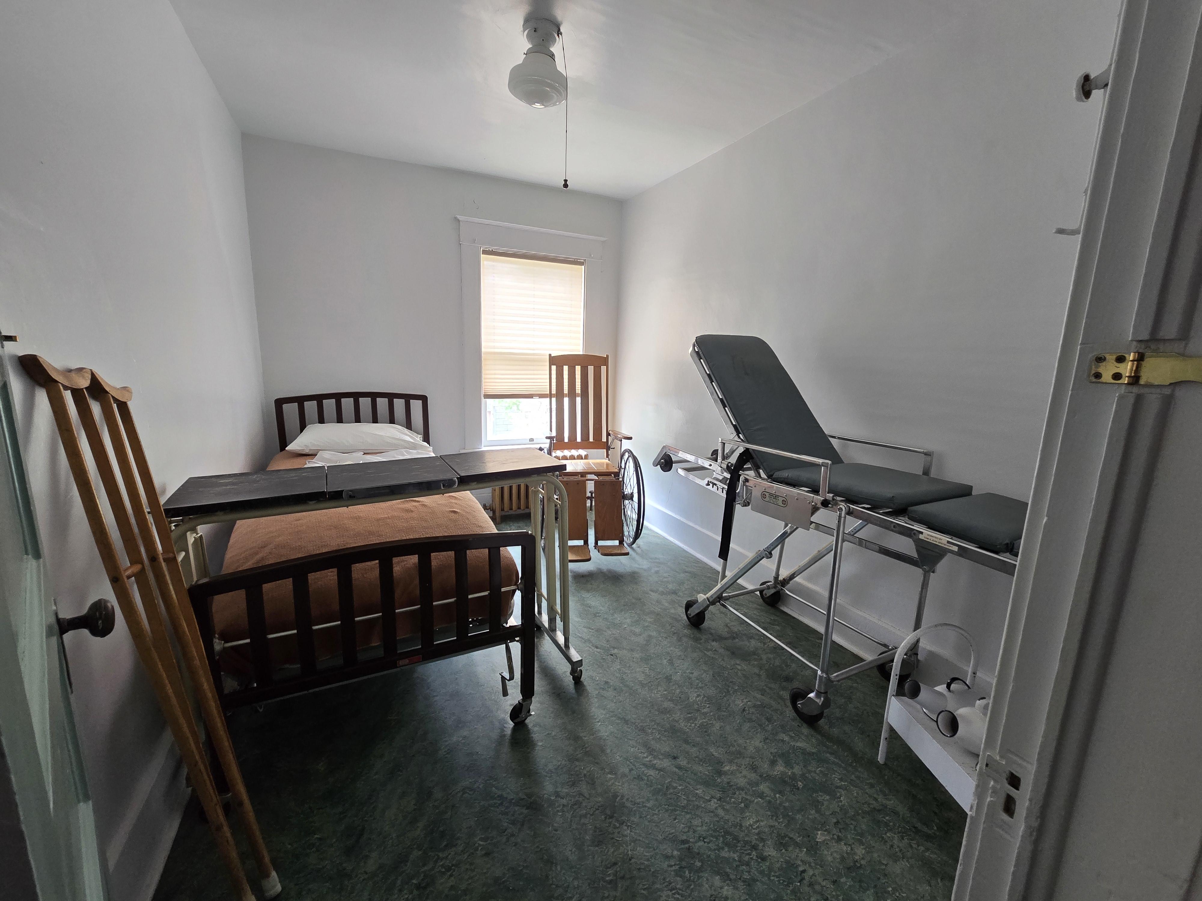 Museum Patient Room, wood crutches, wooden wheel chair, wooden patient bed with orange bedspread, brown and white metal meal tray, chrome metal stretcher with dark gray cushions, white metal tray with white metal urinals